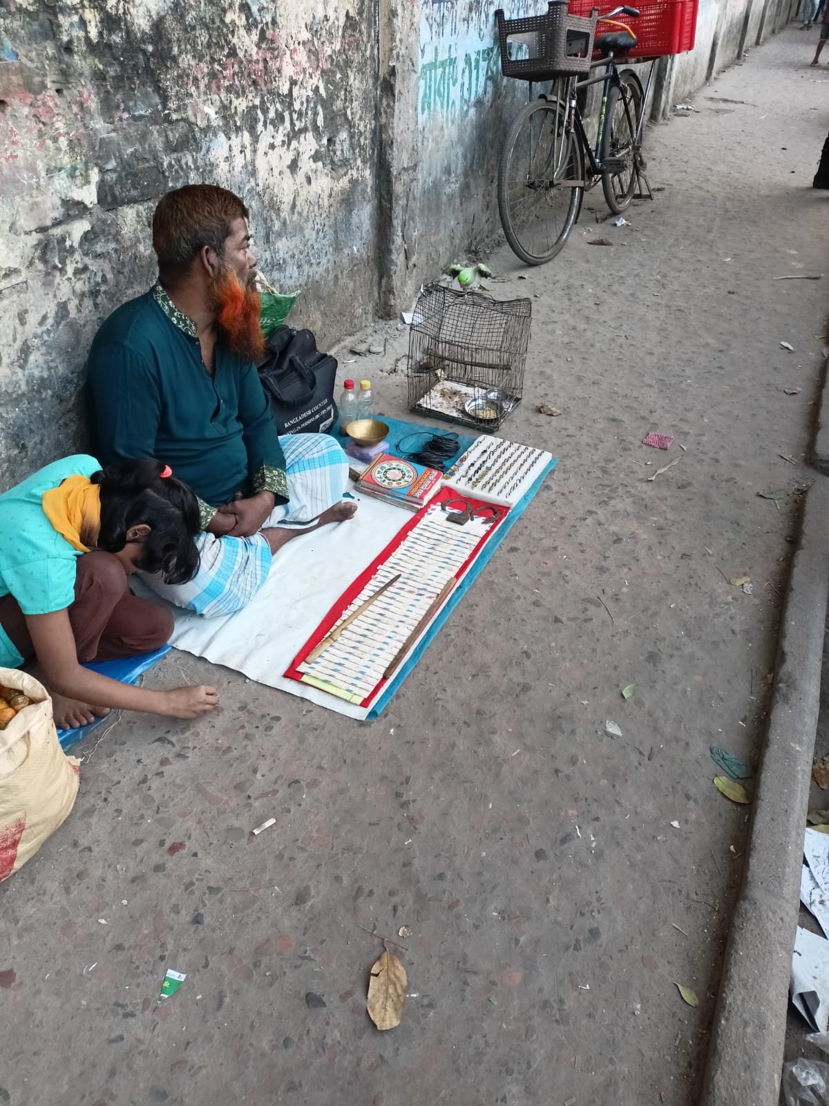 An adult and a child seated beside printed papers and small arranged objects on a pavement next to a weathered wall